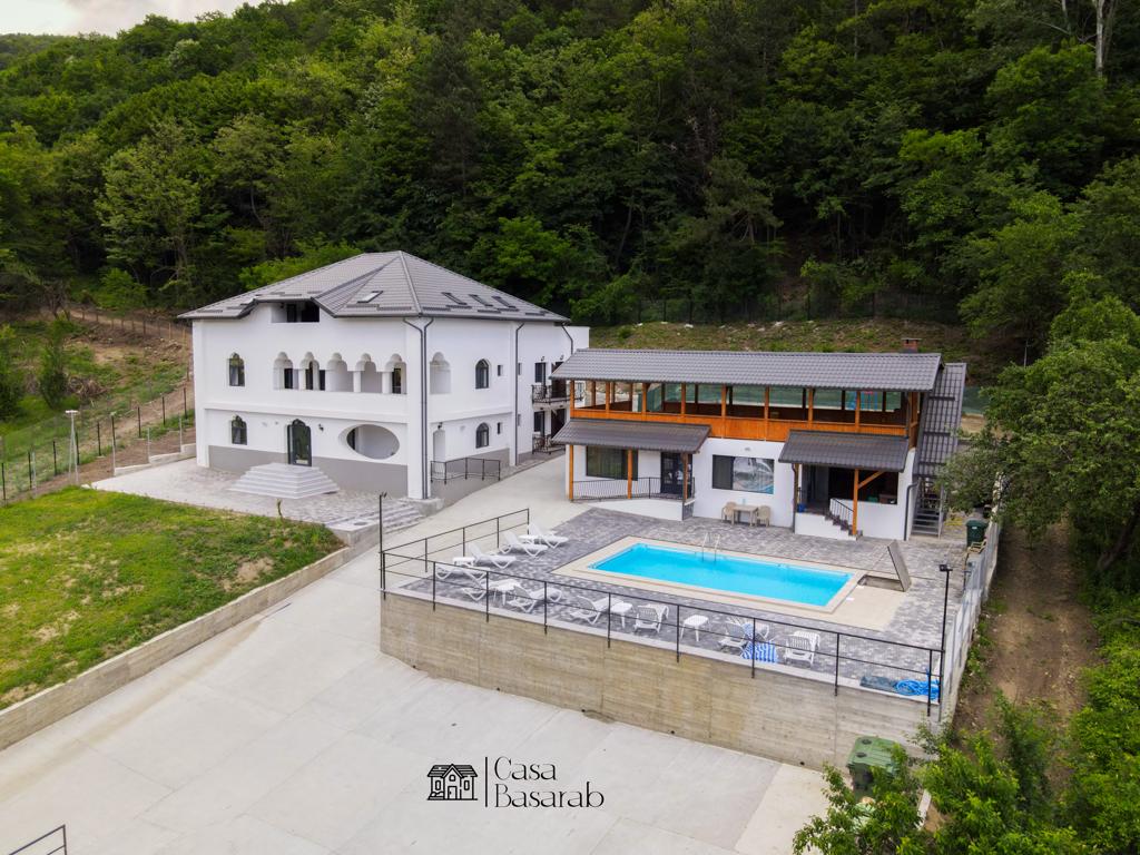 A serene view of two distinct buildings is presented, with a charming white structure featuring arched windows and a dark roof. A modern building with wooden accents encompasses a pool area, surrounded by lounge chairs. Lush greenery forms a natural backdrop.