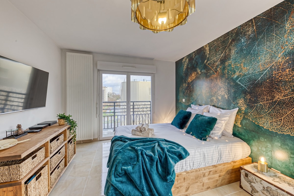 A spacious sleeping area features a large bed adorned with soft pillows and a plush teal throw. A striking wall with leaf motifs provides a bold backdrop. A wooden storage unit holds decorative items, while a large window allows natural light to fill the room.