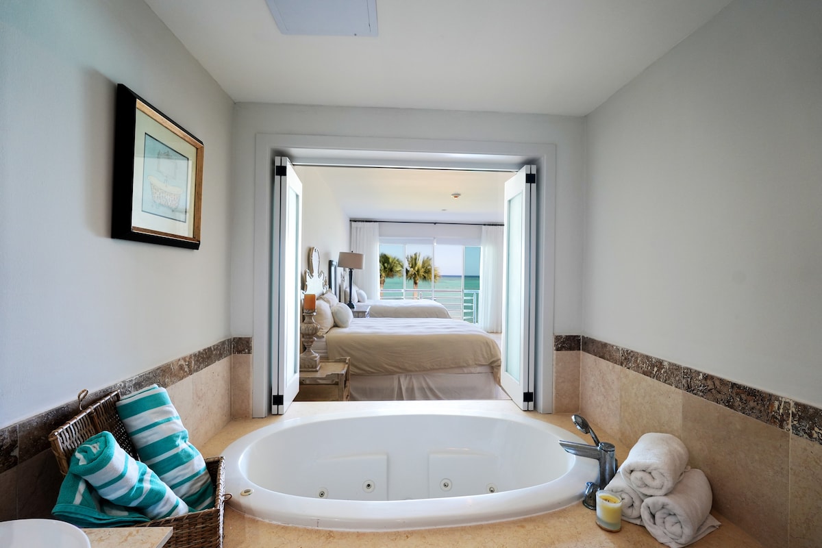 A spacious bathroom features a built-in jacuzzi surrounded by marble accents. Soft towels are neatly arranged beside the tub, and a basket holds additional items. Doorways lead to a well-lit bedroom area with an ocean view and two queen size beds visible through the open doors.