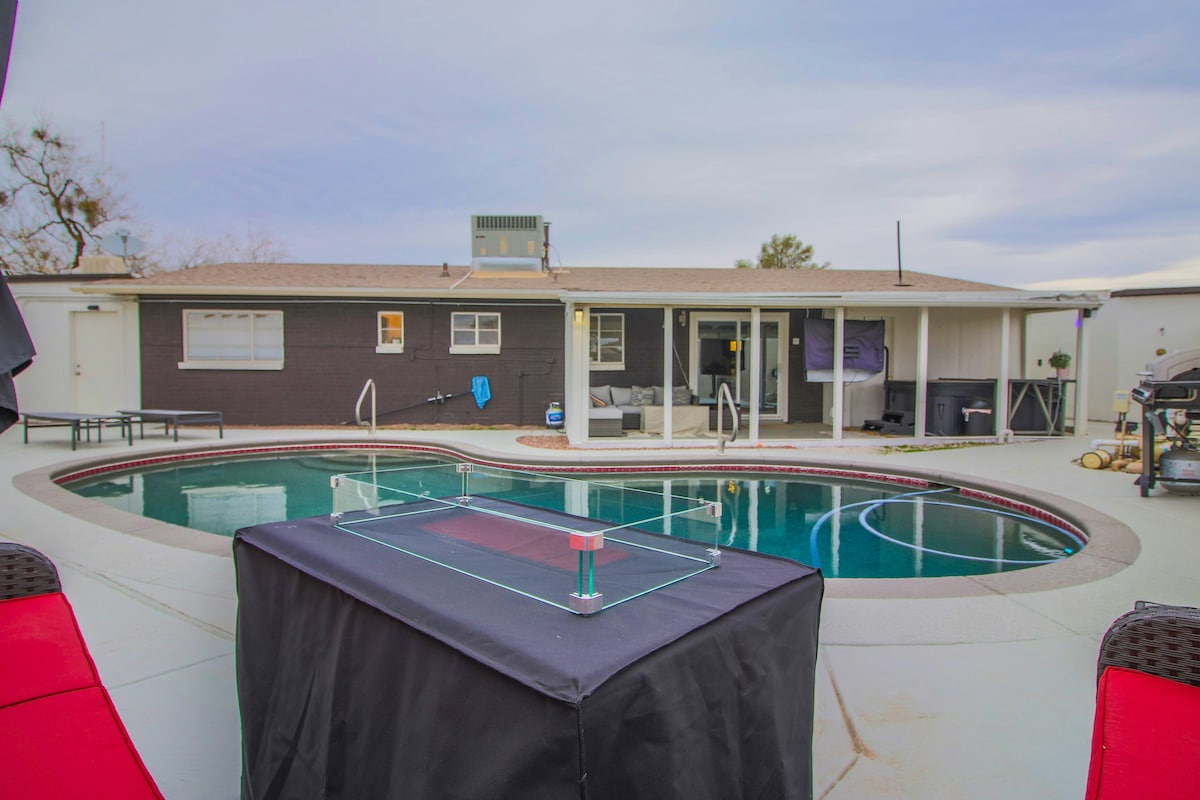 A spacious outdoor area features a sparkling pool surrounded by a sun deck. Comfortable lounge chairs are arranged nearby, with a table in the foreground. The house is visible in the background, showcasing large windows and an inviting patio space.