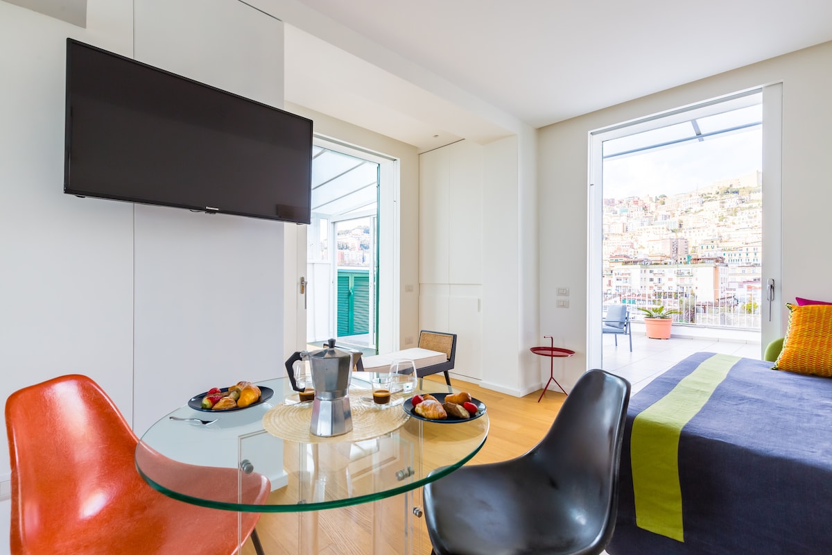 A modern dining area is featured with a glass table set for two, adorned with plates of pastries and coffee cups. A spacious bed is positioned in the background, alongside large windows that offer a view of the vibrant cityscape.
