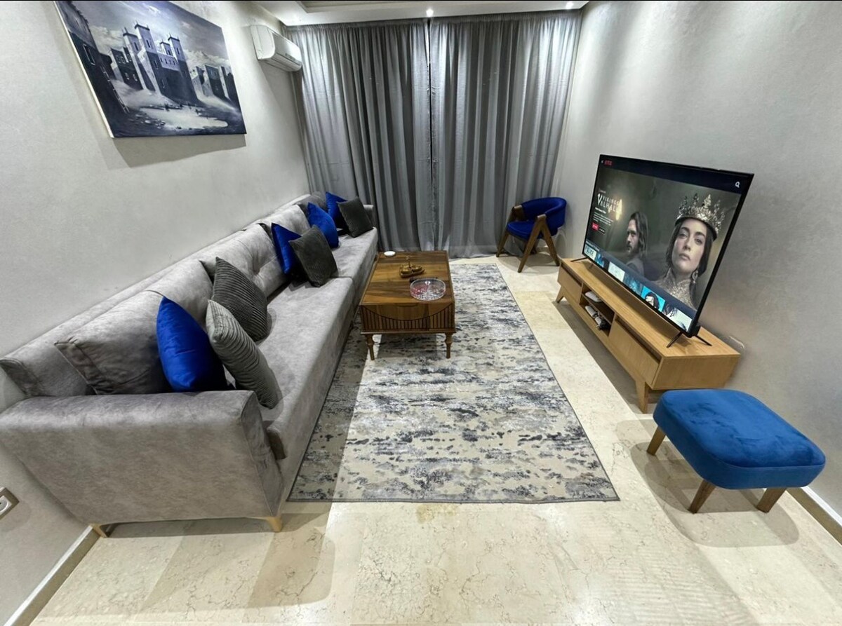 A modern living space features a comfortable gray sofa adorned with blue and patterned cushions. A stylish coffee table sits on a patterned rug, complementing the room's neutral tones. A large television is mounted on a light wood console, and curtains frame a window, allowing natural light to filter in.