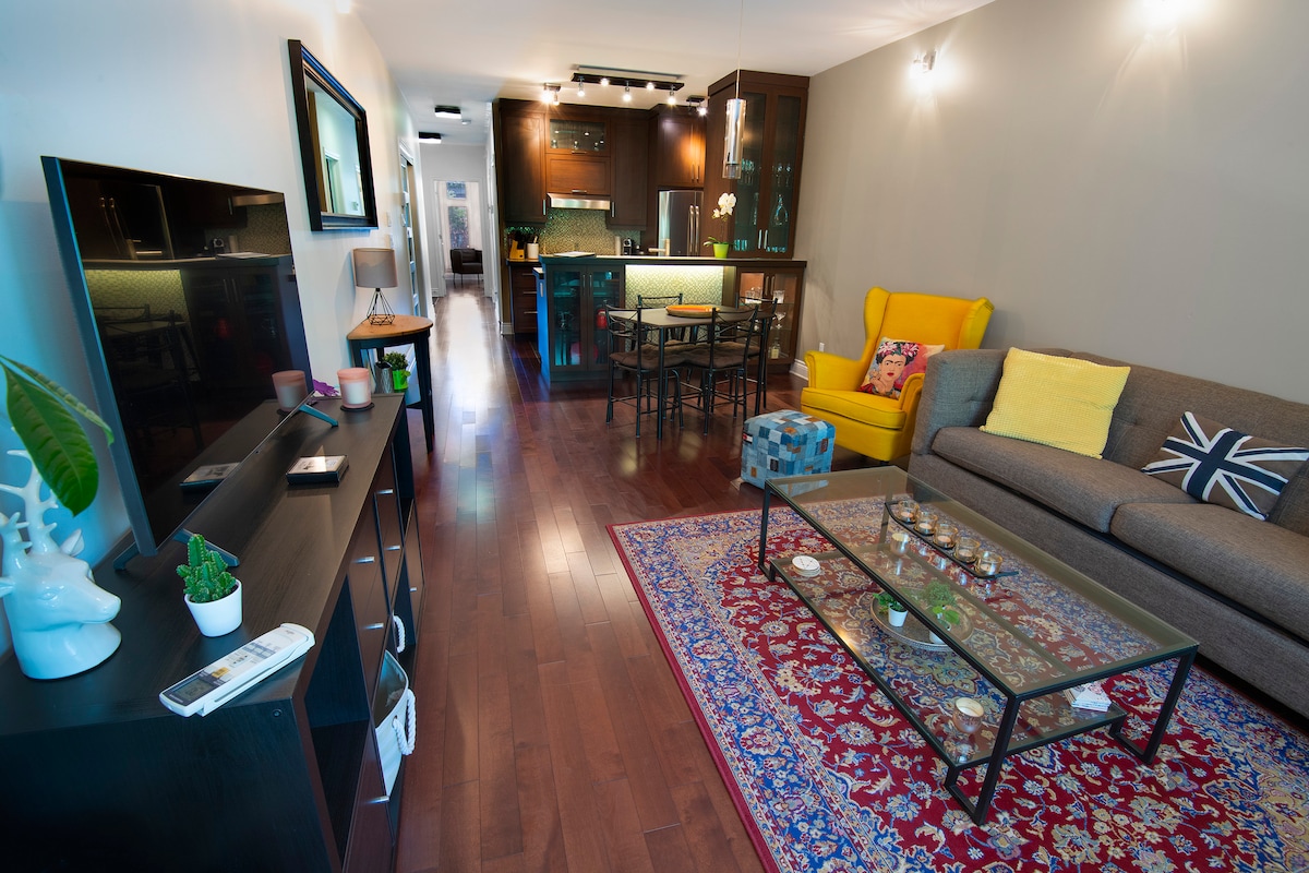 A cozy living area features a sectional sofa and a bright armchair, complemented by a decorative area rug. A coffee table with decorative items sits at the center, and a dining area with a round table is visible in the background, leading to a modern kitchen.