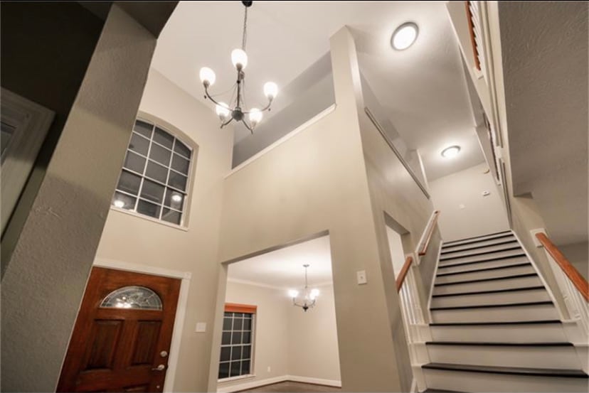 A spacious entryway features a high ceiling illuminated by a modern chandelier. A staircase with a wooden handrail leads to the upper level. A large window allows natural light to fill the area, enhancing the neutral color palette of the walls.