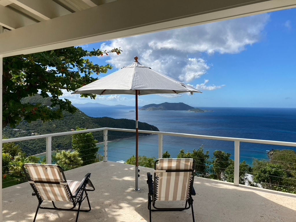 A spacious terrace features two striped lounge chairs placed under a large white umbrella. The view showcases a serene ocean landscape with distant islands and lush greenery, framed by the bright blue sky with fluffy white clouds.