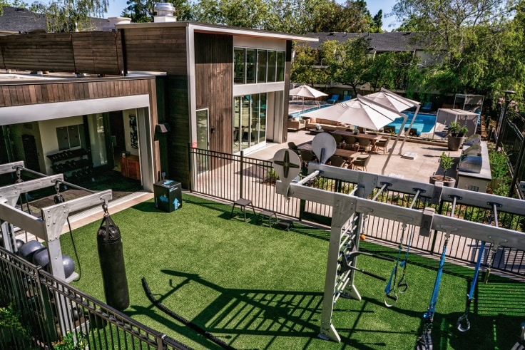 An outdoor fitness area features various workout equipment, including a punching bag and suspension trainers, situated on textured green turf. The space overlooks a swimming pool surrounded by lounge chairs and umbrellas, with stylish wooden structures providing a modern design aesthetic.