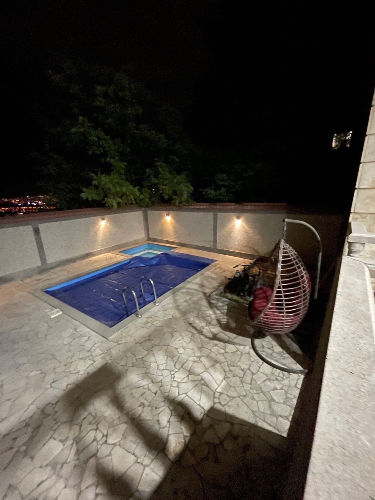 An outdoor area showcases a rectangular swimming pool illuminated softly by built-in lights. A hanging chair is positioned nearby, and the surrounding stone patio features textured tiles. The backdrop includes lush greenery, providing a natural contrast to the modern design.
