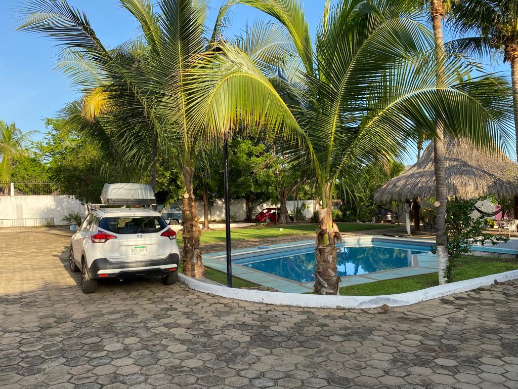 A spacious outdoor area features a communal pool surrounded by lush palm trees. A car is parked nearby on a stone surface, with a palapa offering shaded seating. The well-maintained grounds provide a relaxing environment amidst tropical greenery.