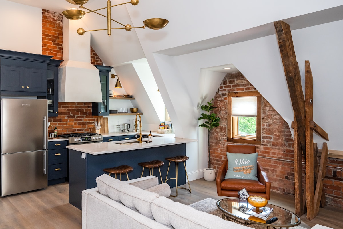 A modern kitchen area features exposed brick walls and a central island with bar seating. Stainless steel appliances and dark cabinetry are present, complemented by stylish light fixtures. A comfortable seating area includes a light gray sofa and a single brown chair, creating an inviting space.