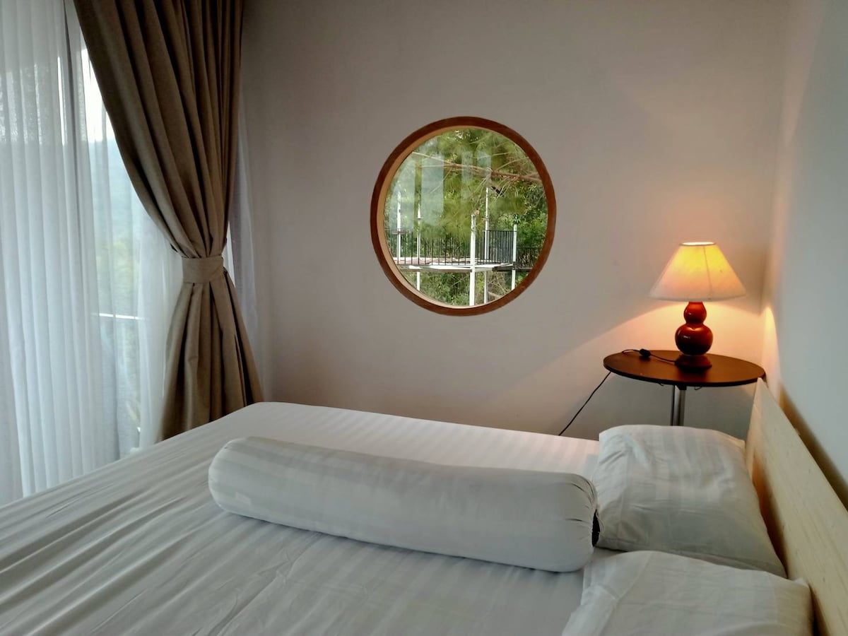 A calming bedroom features a spacious bed with a rolled bolster pillow. Soft, light curtains frame a round window that offers a view of greenery outside. A bedside table holds a simple lamp, providing gentle illumination.