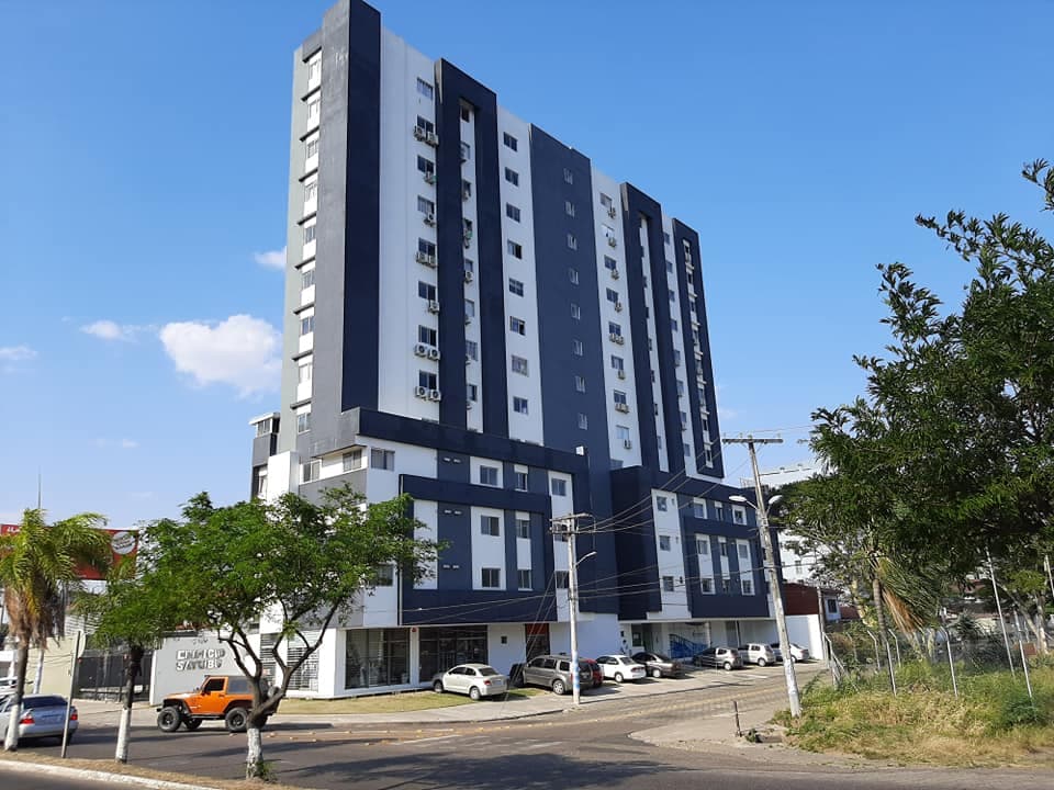 The modern condominium building features a sleek design with multiple levels, painted in contrasting shades of blue and white. Large windows are visible, allowing natural light to brighten the interior spaces. Surrounding greenery and parked vehicles are also present.