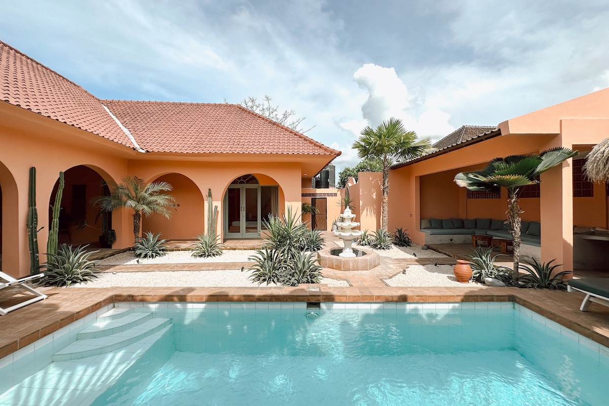 A spacious courtyard features a clear swimming pool surrounded by tropical plants and stone pathways. The villa showcases Spanish-inspired architecture with arched doorways and a small decorative fountain. Comfortable seating is provided under a shaded area adjacent to the courtyard.