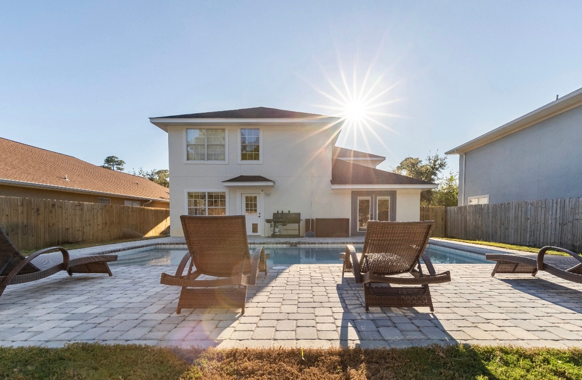 A private outdoor pool area is shown, featuring multiple lounge chairs arranged around the water. The two-story home is visible in the background, with sunlight casting a warm glow over the scene. A fenced yard surrounds the space, enhancing privacy.