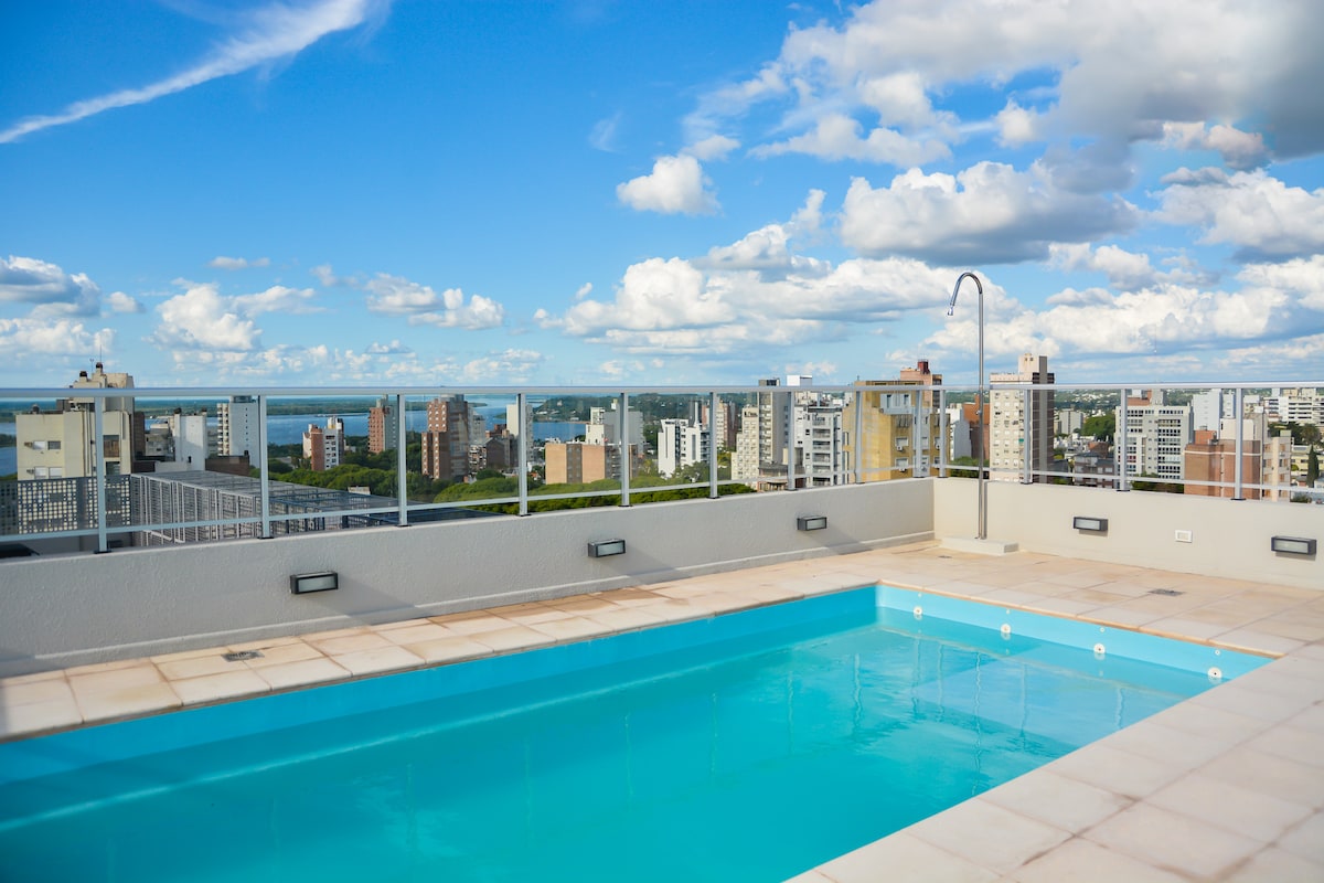 A rooftop terrace features a refreshing swimming pool surrounded by panoramic city views. Clear blue skies with scattered clouds create a serene atmosphere. The pool area is designed for relaxation, offering a spacious environment on a sunny day.