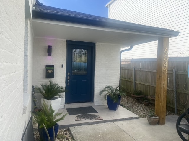 A welcoming entrance features a blue front door with decorative glass and a light fixture, framed by white brick walls. Potted plants add greenery alongside the pathway, which leads to a small mat and a mailbox mounted on the wall.