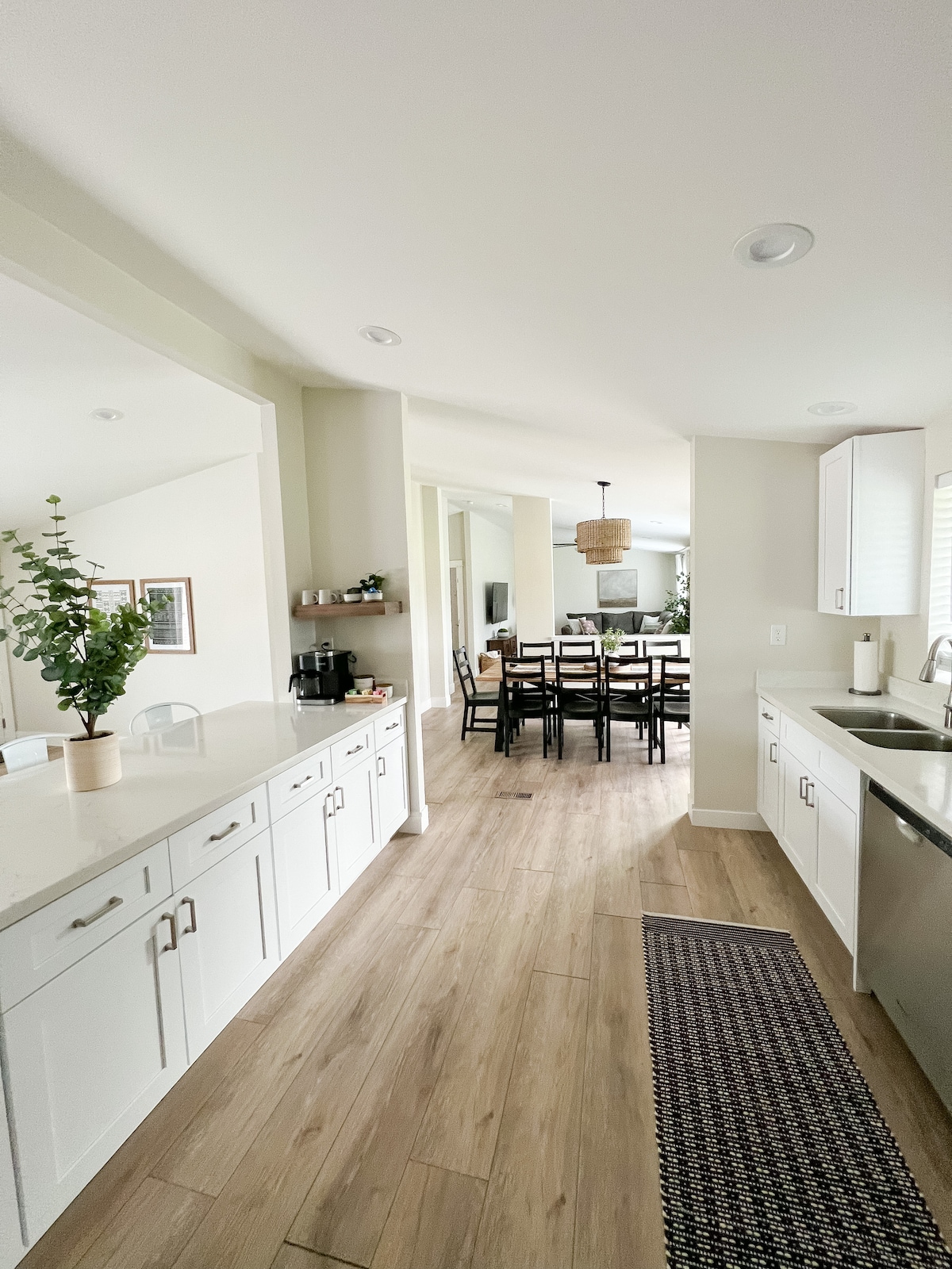 A modern kitchen showcases white cabinetry with sleek hardware, complemented by a light countertop. Natural light filters through the nearby window, illuminating the adjacent dining area with a spacious table and minimalist chairs. A potted plant adds a touch of greenery to the scene.