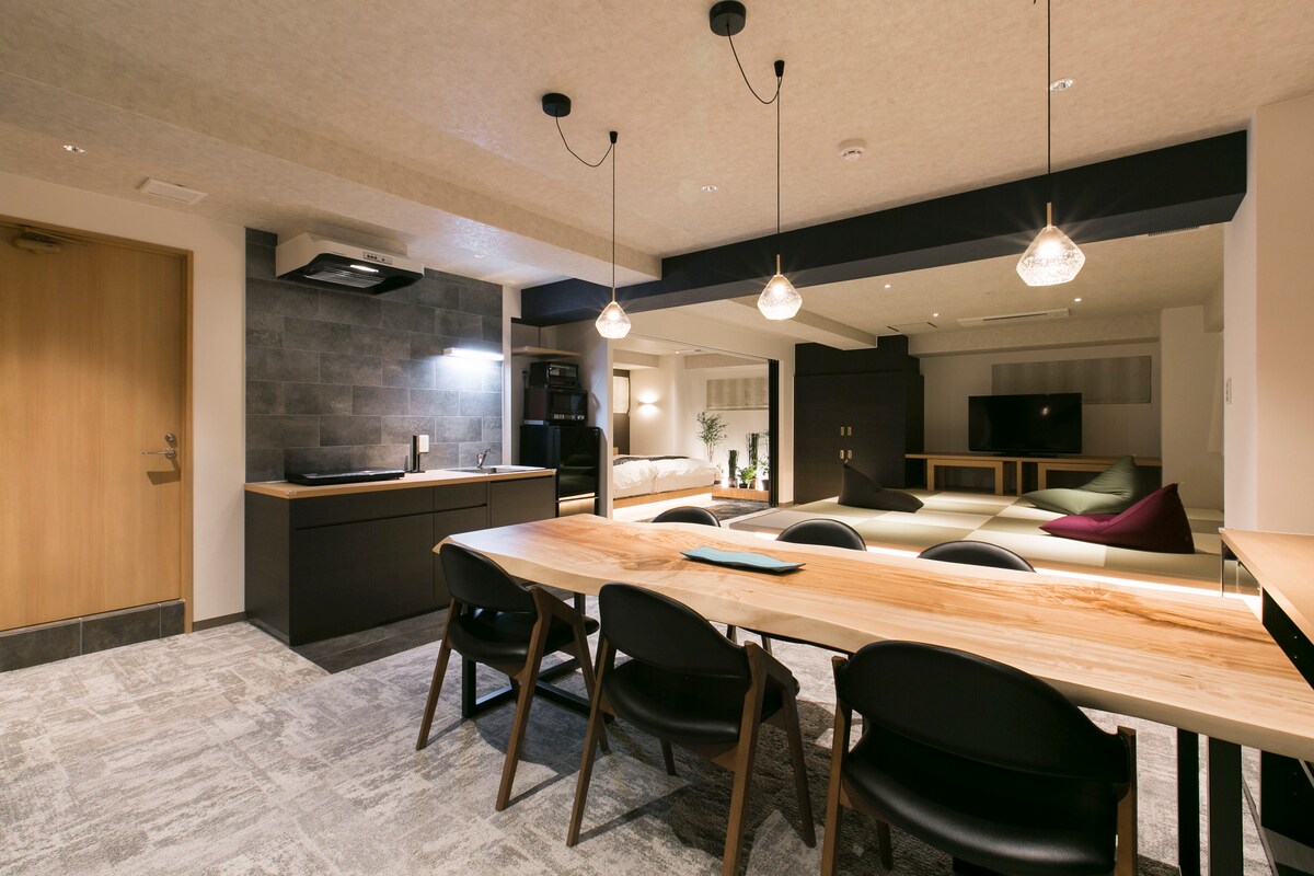 An open living area features a long dining table with black chairs, surrounded by warm, natural wood accents. A kitchenette with modern appliances is visible, along with a cozy seating area showcasing soft, layered cushions. Soft lighting emphasizes the spaciousness and functionality of the area.