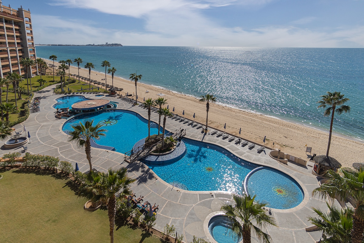 The resort's pools are outlined by palm trees and lounges, leading to a sandy beach. The sparkling blue waters of the ocean are visible in the background, reflecting sunlight under a clear sky. Two jacuzzis are positioned near the pool area.