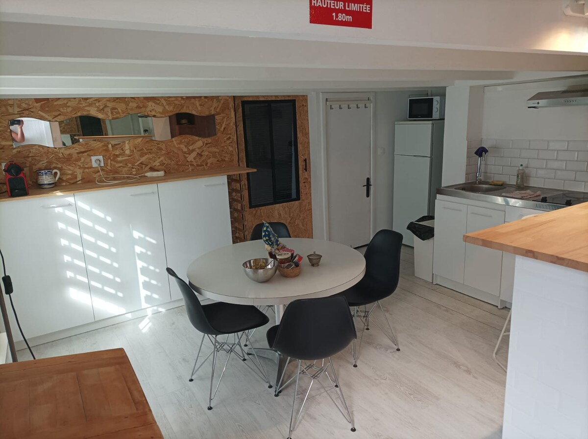 The kitchen area features a round dining table surrounded by black chairs. White cabinets and a light countertop create an airy feel. A small refrigerator is visible, alongside a modern stove and sink. Natural light filters through the windows, enhancing the space's functionality.