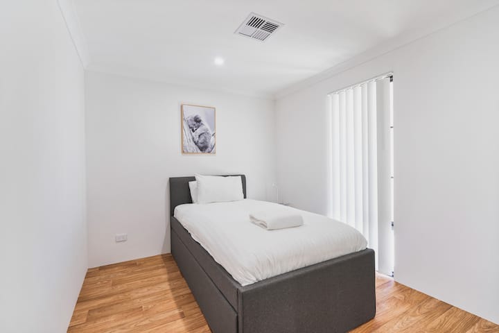 El dormitorio cuenta con una cómoda cama individual con sábanas blancas nuevas y una almohada suave. La luz natural entra a través de persianas verticales, y la habitación tiene pisos de madera y una decoración sencilla y moderna con una pieza de arte de pared enmarcada.