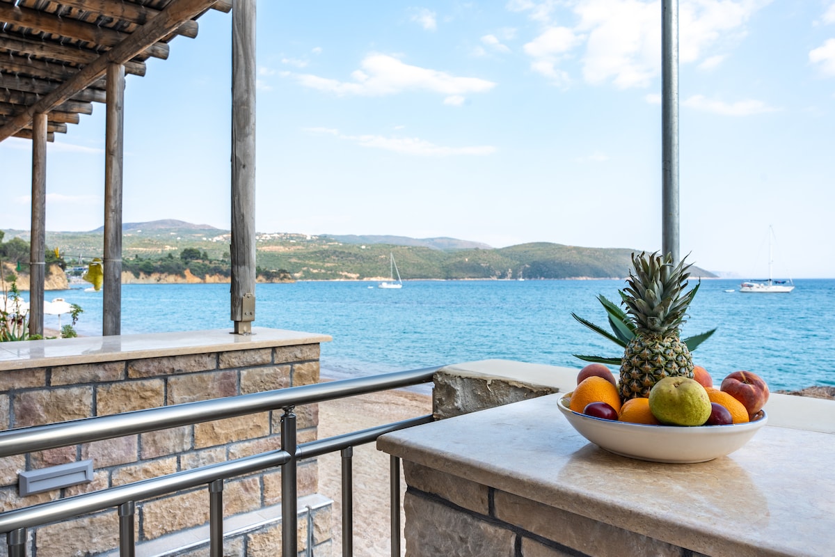 An outdoor seating area features a stone countertop with a bowl of assorted fruits, including a pineapple and citrus. In the background, boats are visible on the serene blue water, framed by a coastline and distant hills under a clear sky.