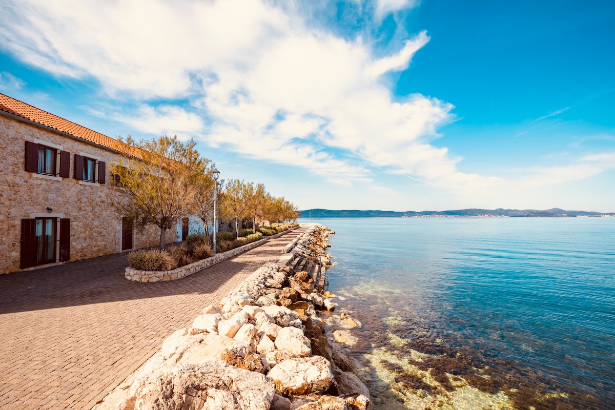 The image captures a serene waterfront scene featuring a rocky shoreline adjacent to calm waters. A path lined with trees runs alongside a stone building, with a clear sky and gentle clouds creating a tranquil atmosphere. The landscape showcases the natural beauty of the coastal area.