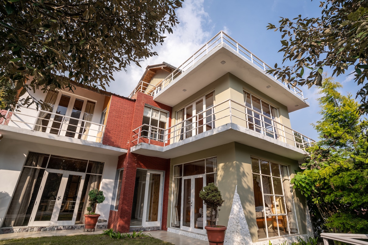 The villa's modern architecture is highlighted by large glass doors and expansive balconies on multiple levels. Well-manicured greenery and potted plants frame the entrance, complementing the bright exterior and inviting ambiance beneath a clear sky.