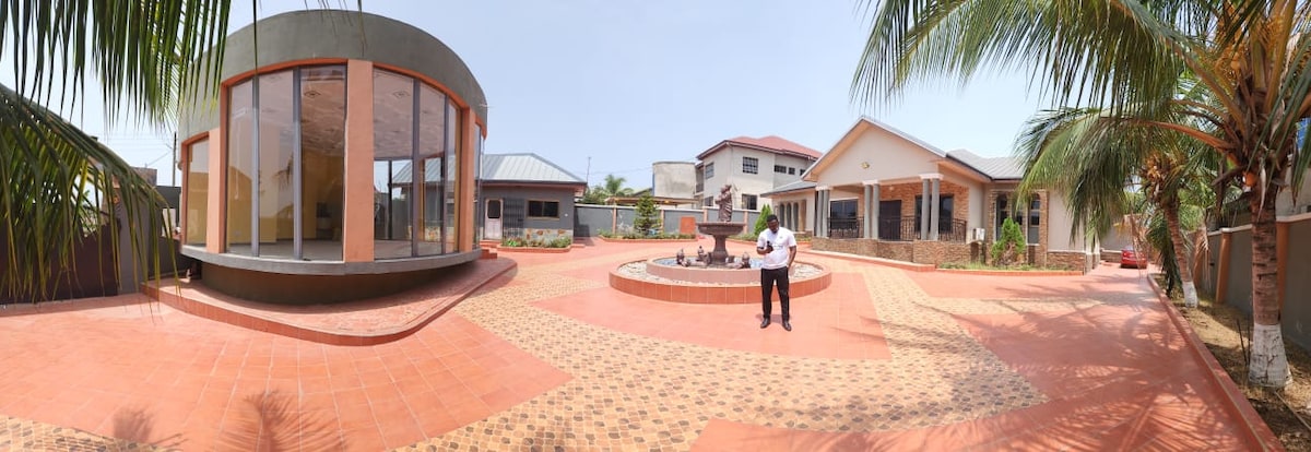 An expansive outdoor area is featured, showcasing a tiled courtyard with a decorative fountain at its center. Surrounding palm trees and residential structures create a relaxed environment, with ample space available for gatherings or leisure activities.