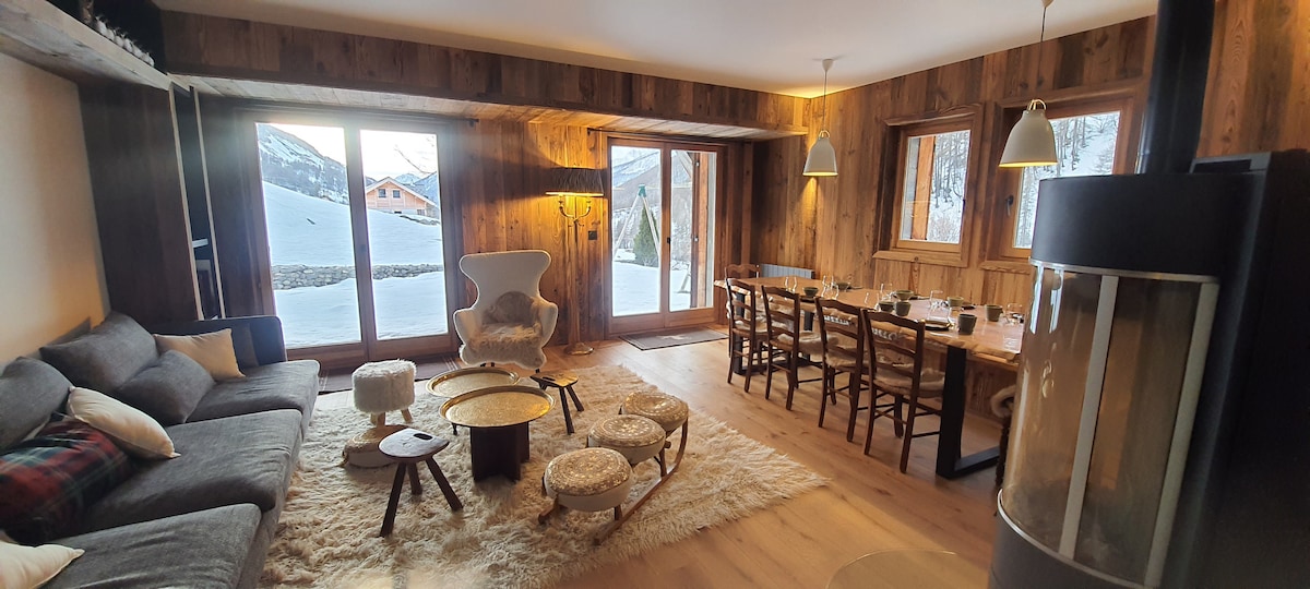 A spacious living area features large glass doors that provide a view of the snowy landscape outside. A comfortable sofa is arranged around a central coffee table, alongside a dining table with wooden chairs. Plush rugs and a fireplace enhance the cozy ambiance.