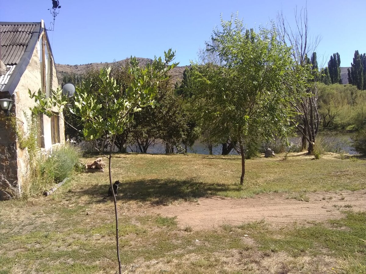 A landscaped area features grassy grounds with a few trees and shrubs, extending toward the Aluminé River. The background showcases a rustic stone building, with mountains and clear blue skies visible above, enhancing the tranquil setting.