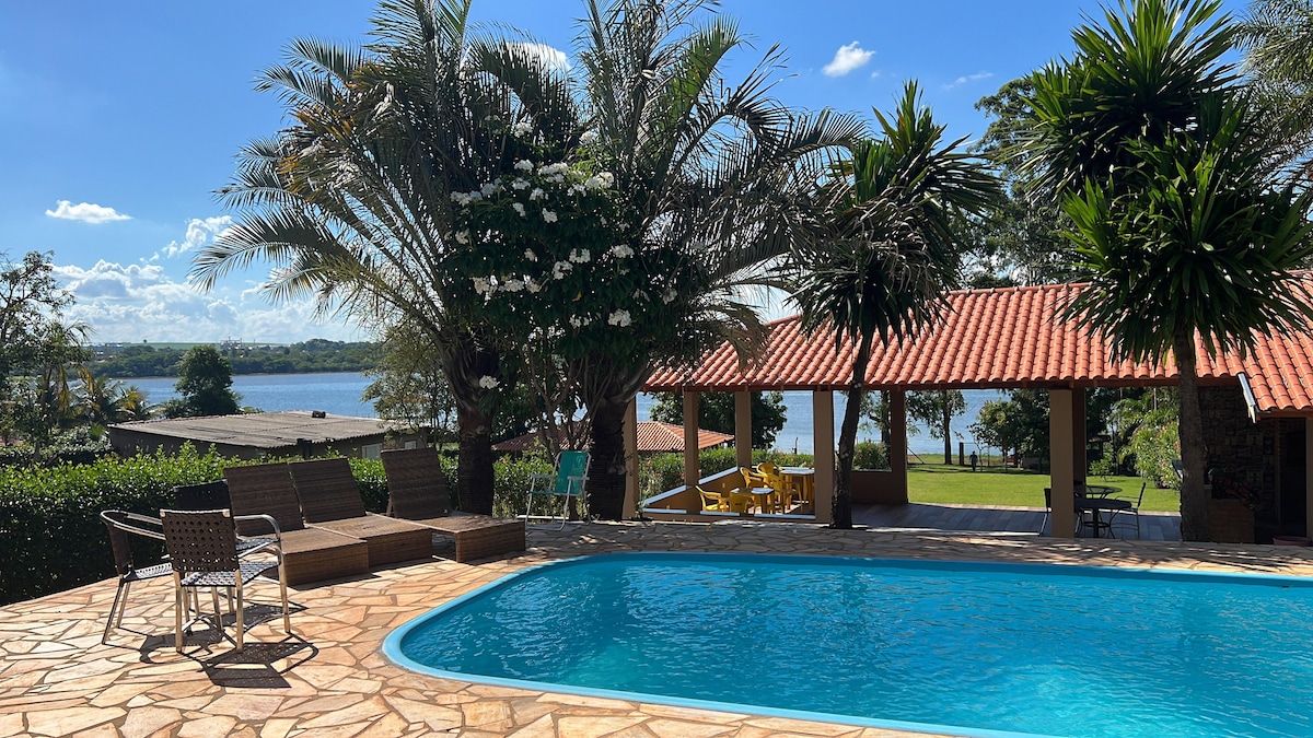 A spacious outdoor area features a heated swimming pool surrounded by lounge chairs. Lush palm trees provide shade, while a picturesque view of the river is visible in the background. A covered seating area with a red-tiled roof is positioned nearby.