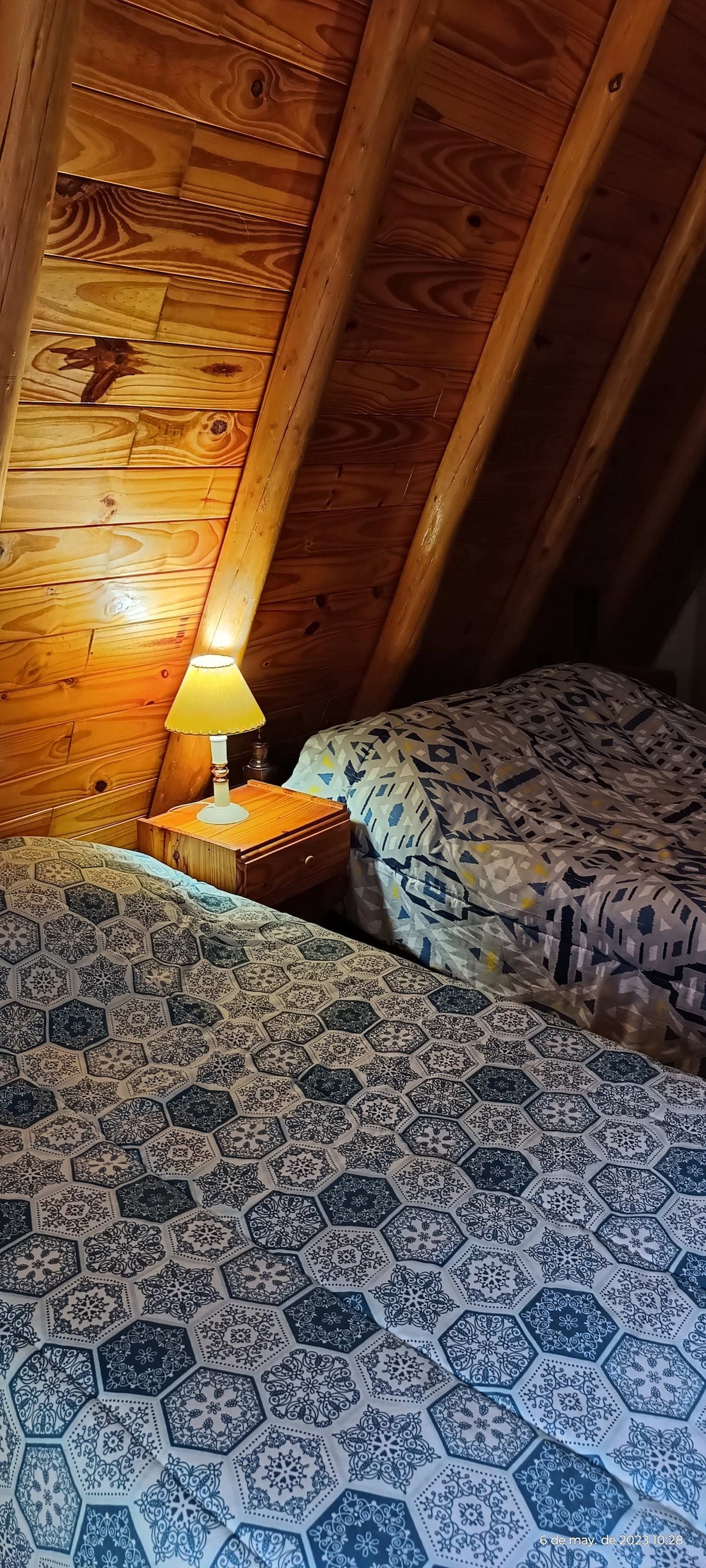 A cozy bedroom is presented with sloped wooden ceilings. Two beds are positioned side by side, each adorned with patterned bedding. A small bedside table holds a lamp, casting a warm light in the space.