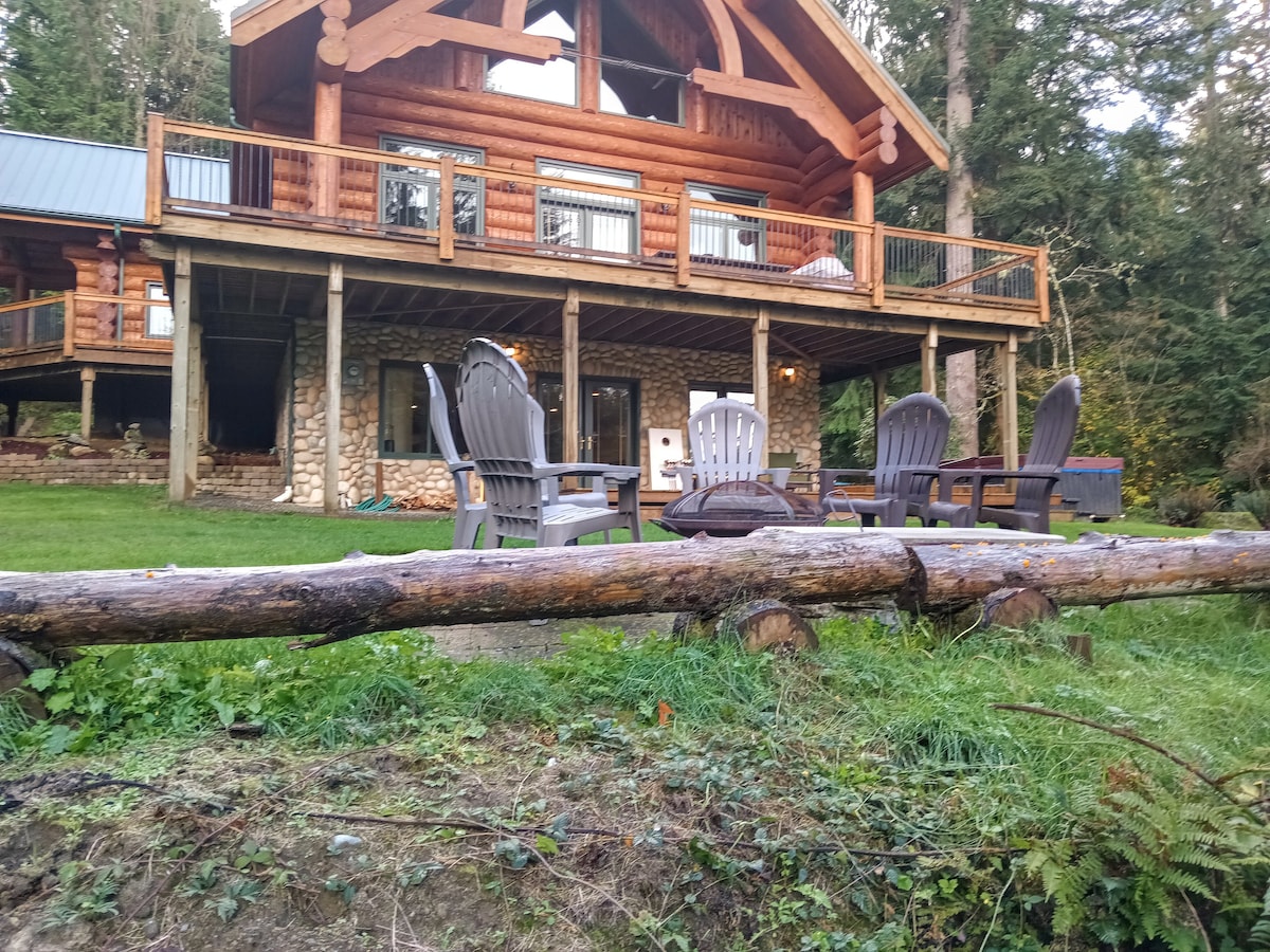 A spacious log house is featured, showcasing a large deck with multiple seating areas. Adirondack chairs are positioned in front of the house, and a rustic log is partially visible in the foreground. Lush greenery surrounds the property, enhancing the natural setting.