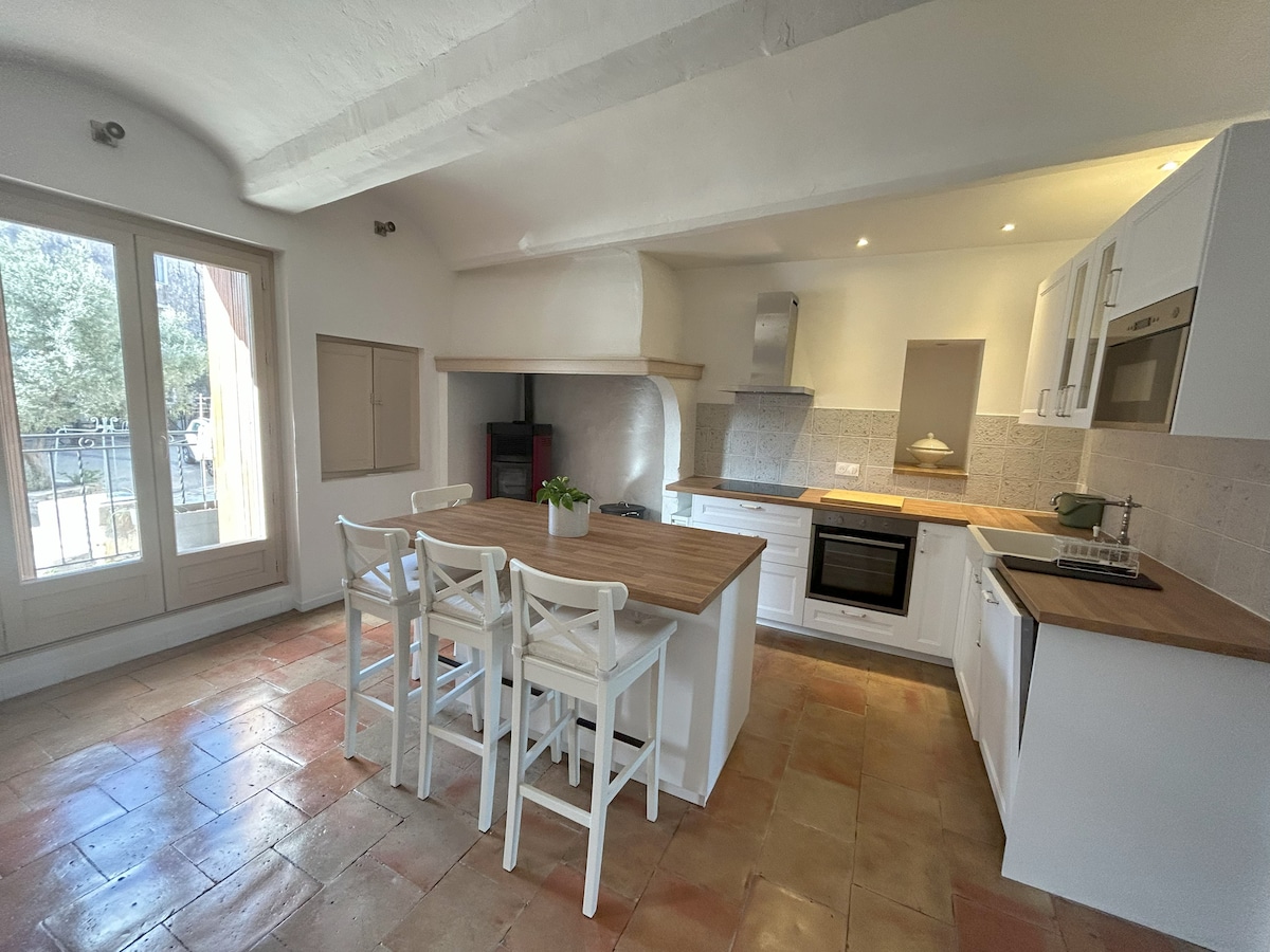 A spacious kitchen is presented, featuring a central island with four white chairs. Modern appliances are integrated, including an oven and a hood. Natural light floods in through large windows, illuminating the terracotta floor and highlighting the clean, minimalist design.