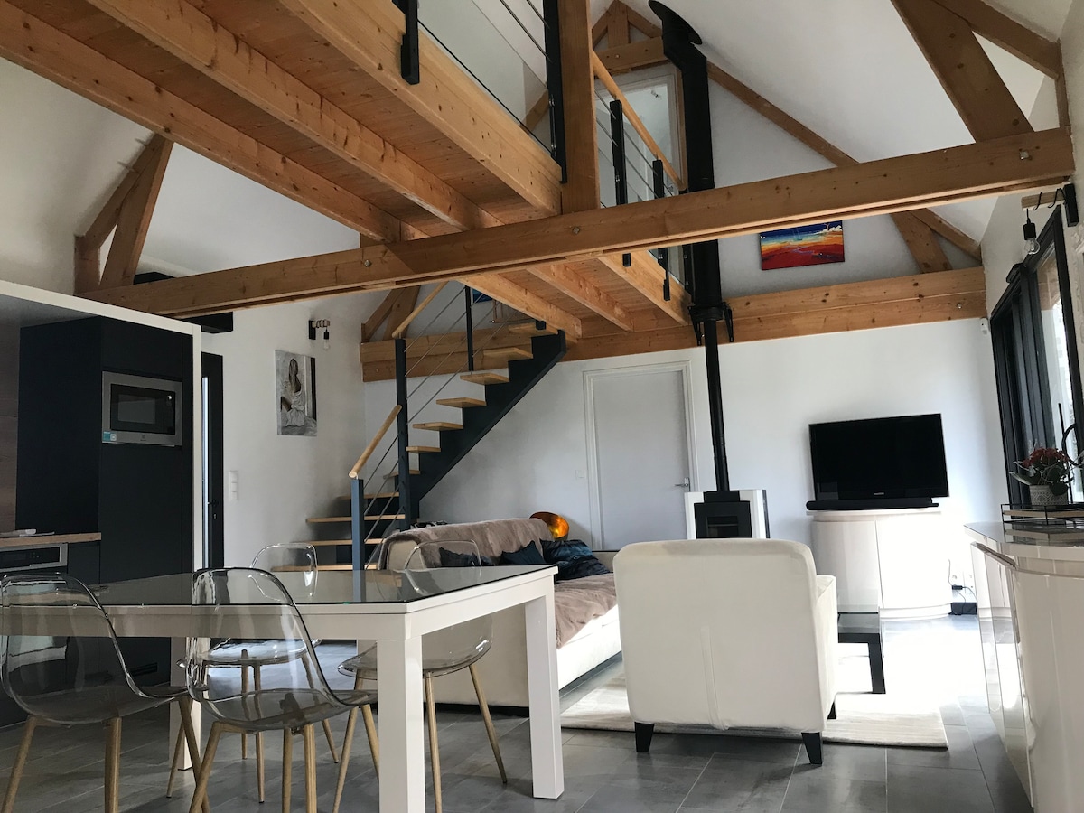An open-concept living area showcases vaulted ceilings supported by wooden beams. A staircase leads to the upper level, where a cozy sleeping area is visible. The space includes a dining table with transparent chairs, a functional kitchen area, and a lounge area featuring a television and a wood stove.