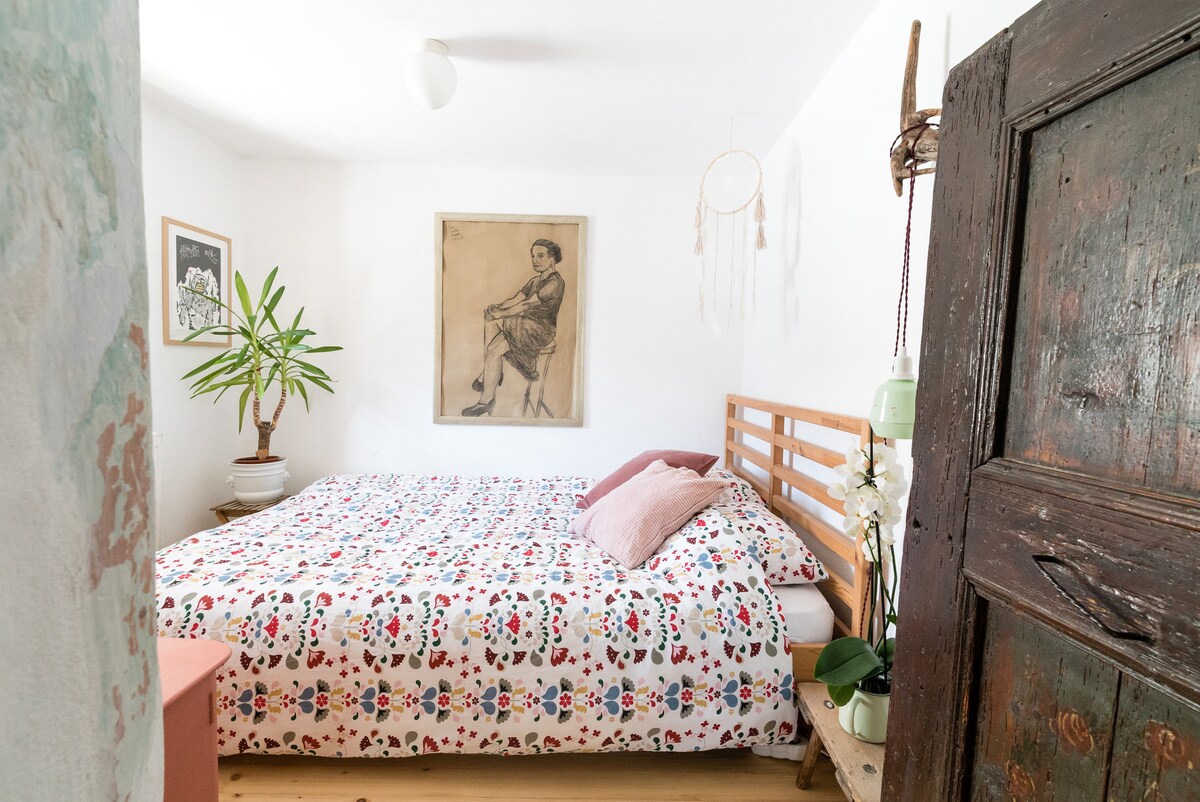 A bright bedroom features a spacious bed adorned with a floral patterned quilt and soft pink pillows. A large art piece hangs on the wall above the bed, complemented by a potted plant in the corner. Natural light illuminates the space, creating a cozy atmosphere.
