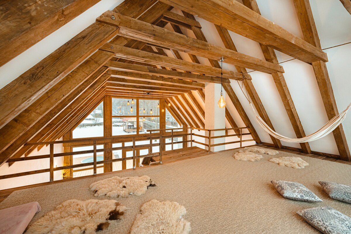 A spacious loft area showcases exposed wooden beams and a cozy atmosphere. Soft textile mats are placed on the floor, accompanied by decorative cushions. A hammock is suspended near large windows, offering views of the surrounding landscape and allowing natural light to illuminate the space.