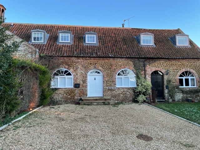 An inviting exterior of a charming brick cottage is showcased, featuring a traditional roof with dormer windows. A clear pathway leads to the front door, surrounded by a landscaped area with greenery. The spacious gravel driveway allows for off-street parking.