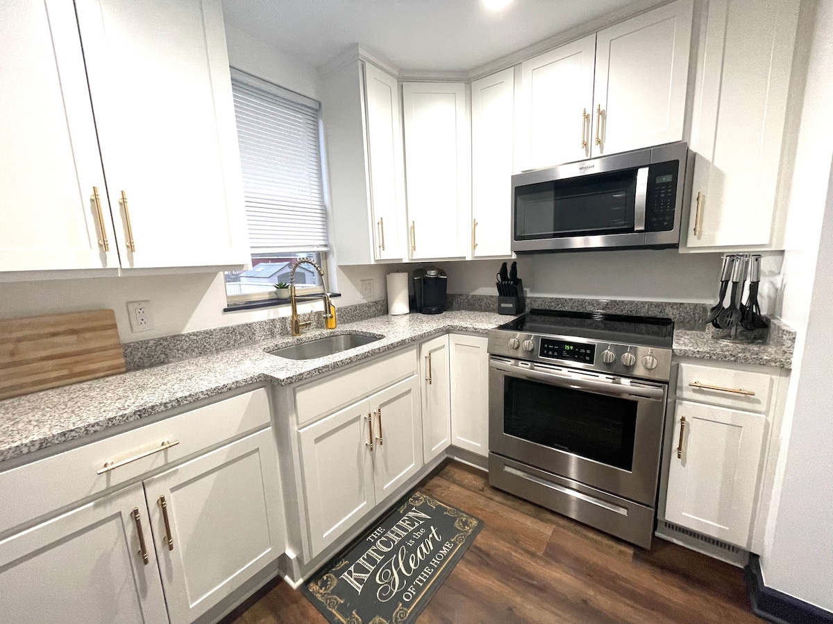 An equipped kitchen features white cabinetry with gold hardware and a granite countertop. Stainless steel appliances, including an oven and microwave, are prominently displayed. A window provides natural light, and flooring in a warm tone complements the overall design, enhancing the kitchen's functionality.