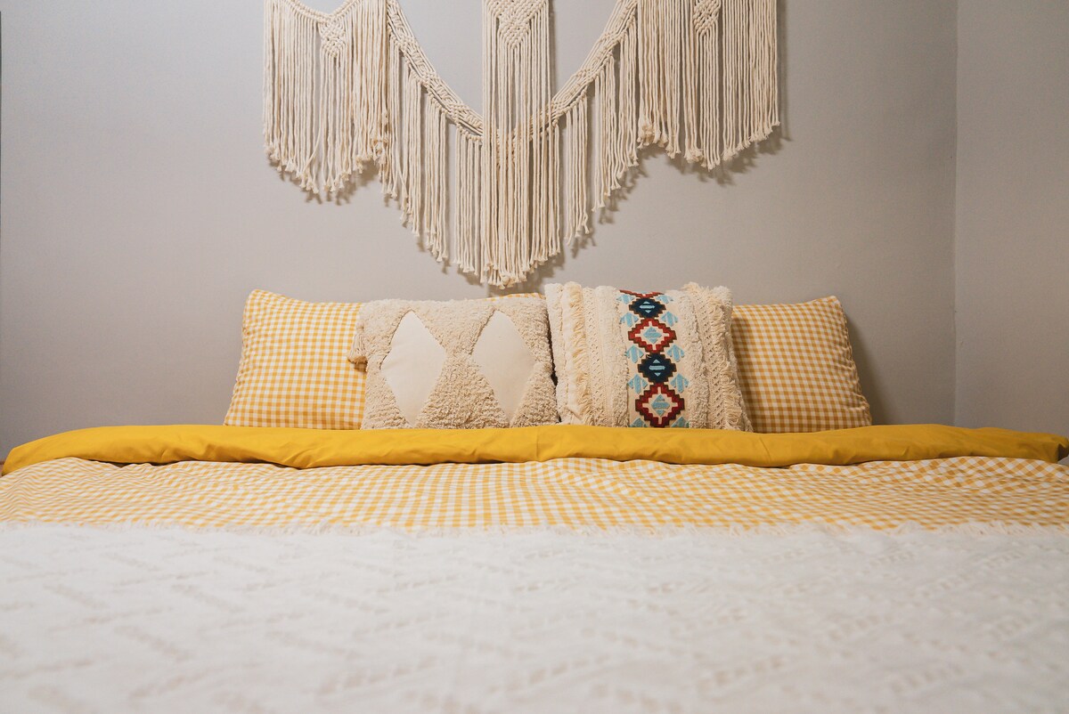 A spacious bed is displayed with a yellow and white checkered duvet cover and decorative pillows. A textured wall hanging adds a touch of character above the bed, contrasting with the soft gray walls.