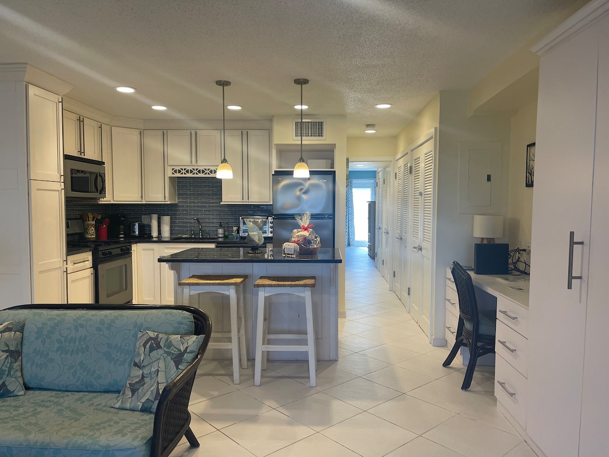 The kitchen area showcases white cabinetry and stainless steel appliances, including a full-size stove and refrigerator. A breakfast bar with two highboy chairs is present. A living area includes a sofa with patterned cushions. Light tile flooring extends towards a hallway transitioning to additional rooms.