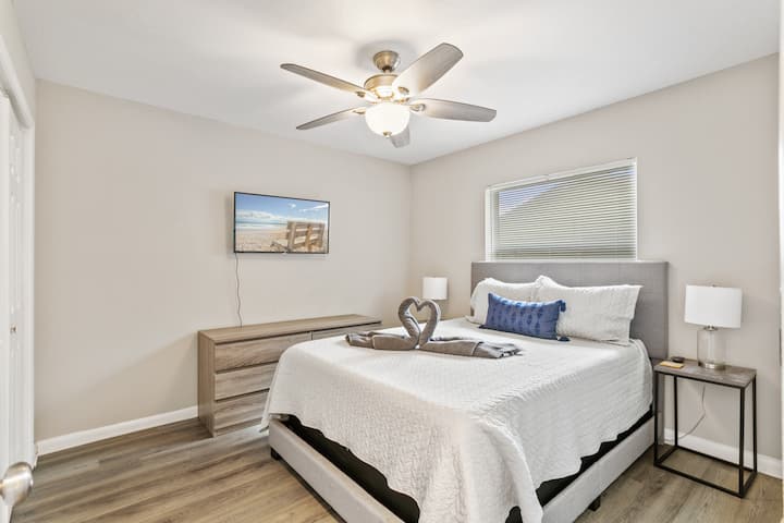 Queen Bedroom ახალი თეთრეულით, საწოლისპირა მაგიდა ლამპით, ვენტილატორით და სმარტ-ტელევიზორით.