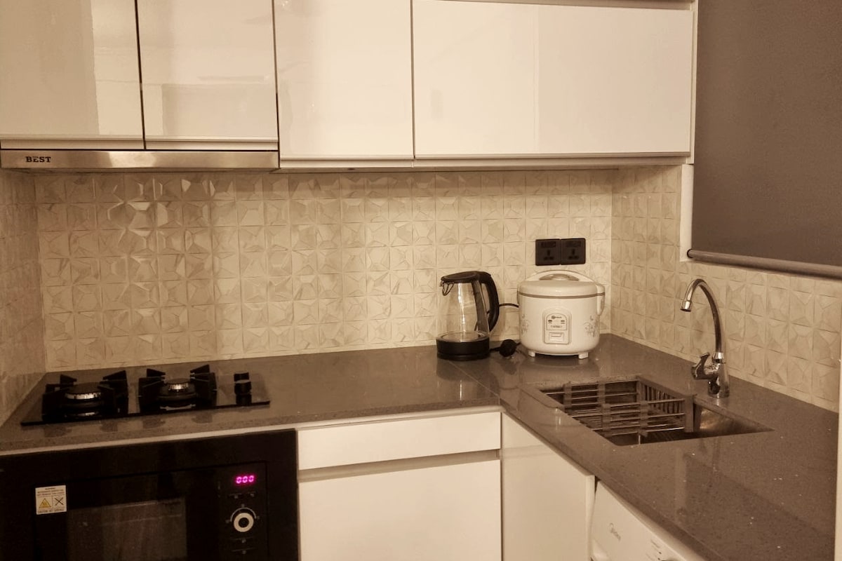 A modern kitchen features sleek white cabinetry and a textured backsplash. Stainless steel appliances, including a gas cooktop and an oven, are complemented by a countertop with a kettle and rice cooker. The chrome faucet sits above a stainless steel sink, enhancing the functionality of the space.