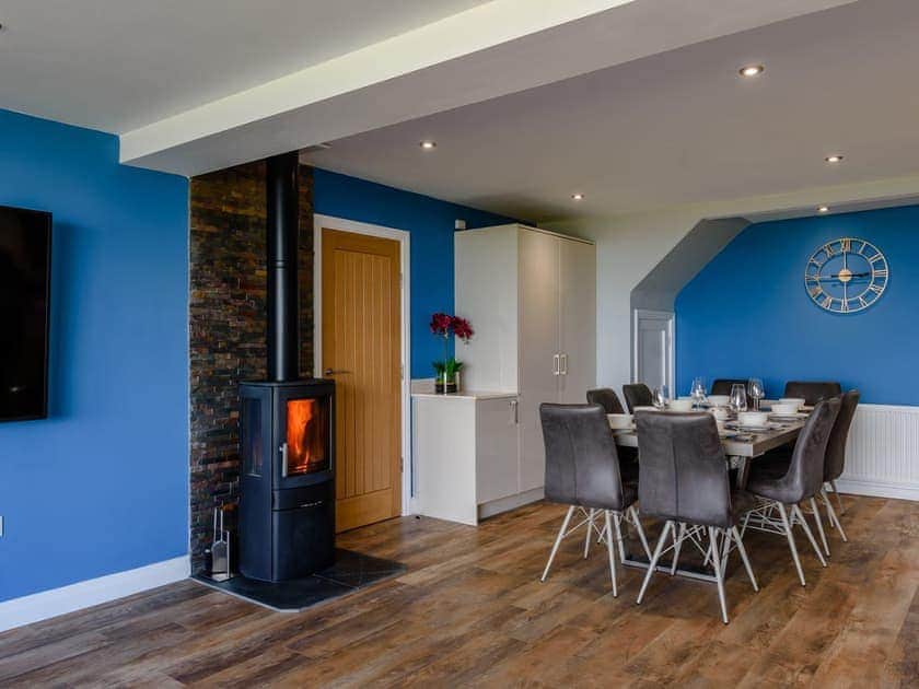 A modern dining area features a spacious table surrounded by six grey chairs, set against a vibrant blue wall. A sleek wood burner adds warmth to the room, while a large television and a decorative wall clock complete the inviting atmosphere.