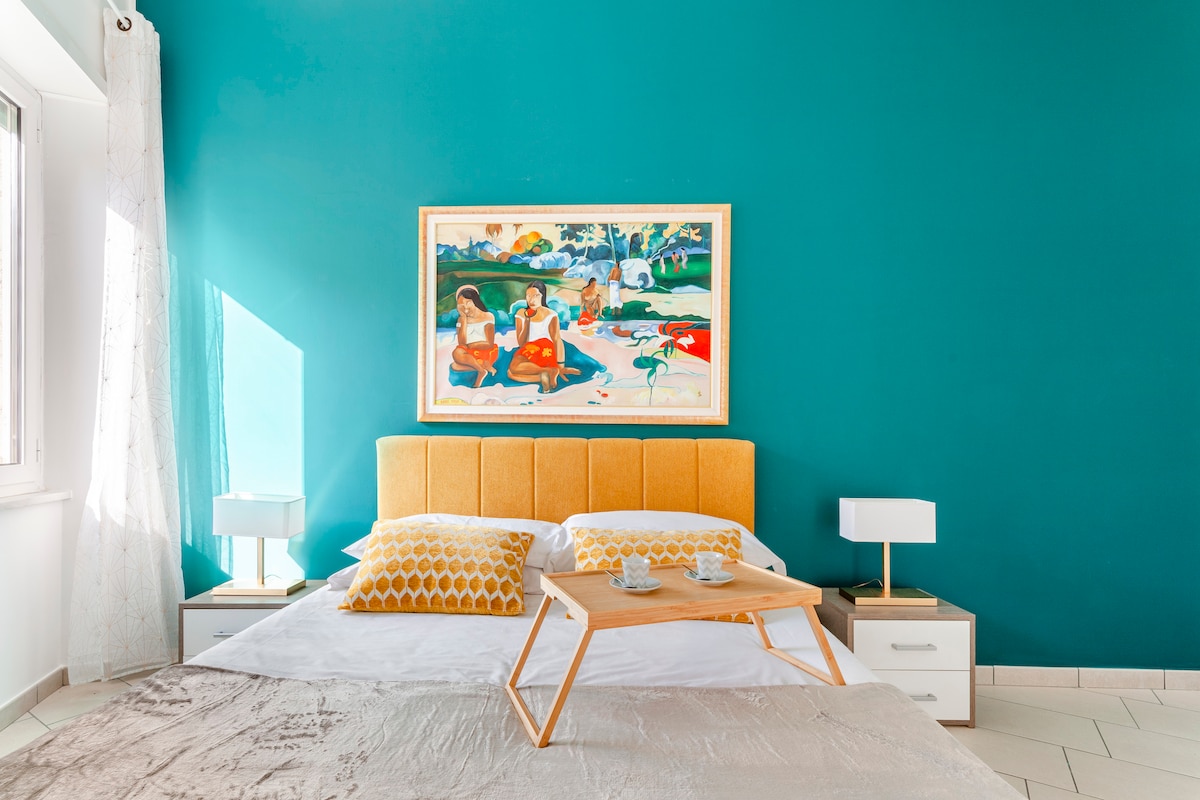 A bright sleeping area features a spacious bed with a yellow headboard and decorative pillows. A painting of three figures is displayed above the bed on a vibrant teal wall. Nightstands with white lamps are positioned on both sides of the bed.