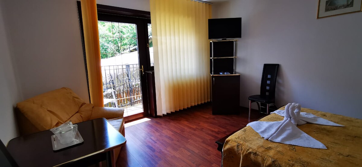 A brightly lit room features a spacious bed with a gold-colored coverlet. A comfortable armchair is positioned next to a small table, while a flat-screen TV is mounted on a shelf. Sheer yellow curtains allow natural light to fill the space.