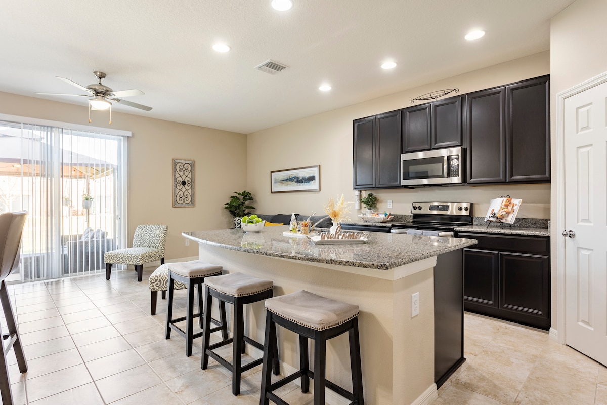 A modern kitchen area features sleek black cabinetry and granite countertops. A central island is accompanied by four padded stools, enhancing the dining space. Natural light is invited through sliding glass doors, leading to an outdoor area, while overhead LED lights offer bright illumination.