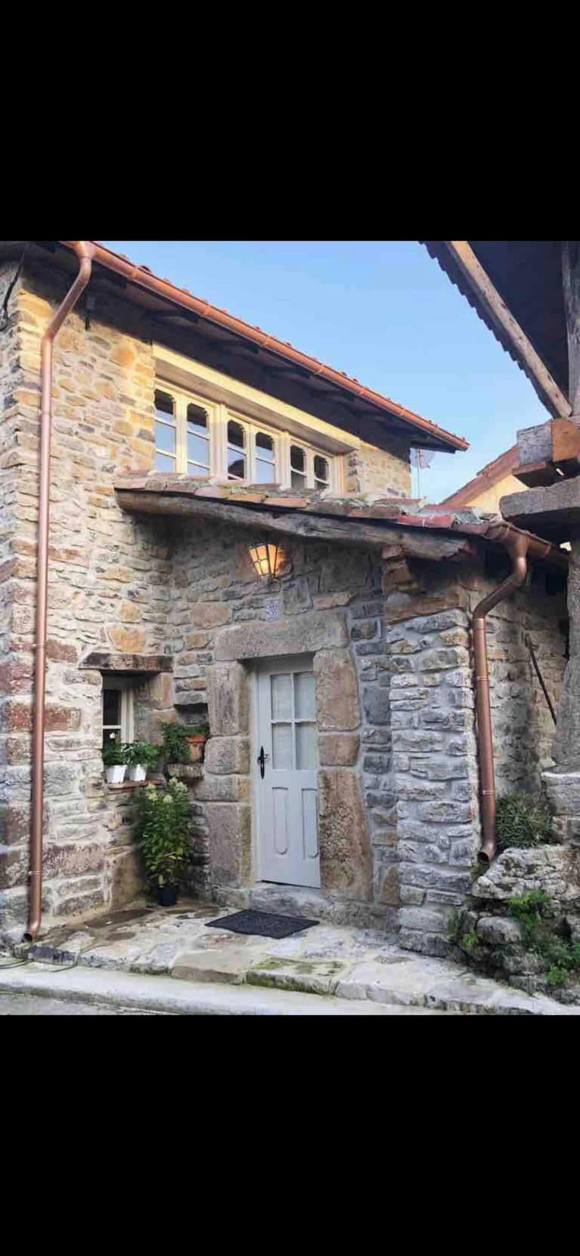 The exterior features a charming stone facade with wooden elements and a lantern above the entrance. Four small windows allow natural light to enter, and greenery is visible beside the front door, contributing to a welcoming appearance.