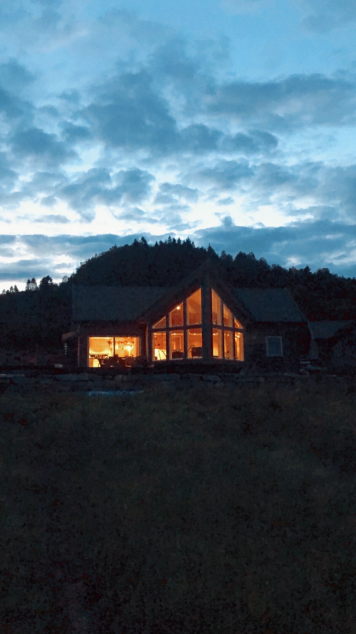 A cabin is illuminated with warm light, showcasing large windows that highlight its inviting architecture. The surroundings feature a serene landscape of trees under a twilight sky, creating a peaceful atmosphere.