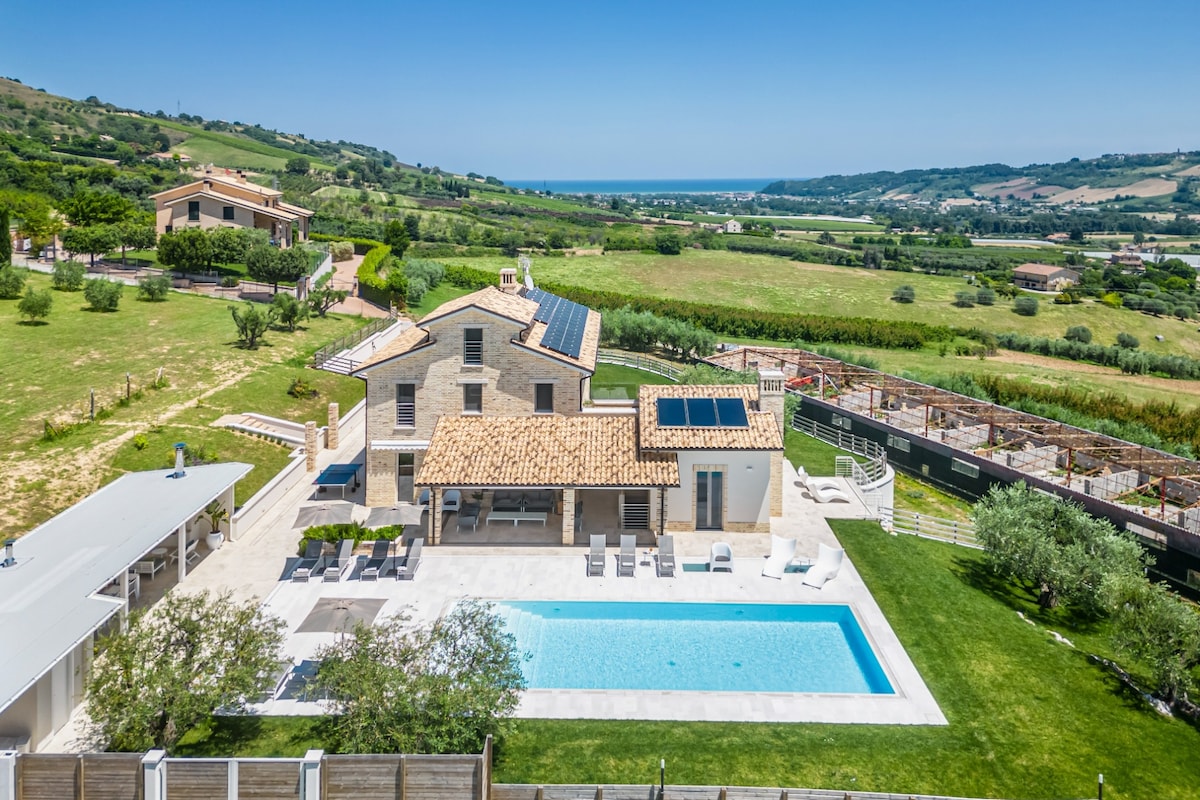 The villa is set against a backdrop of rolling green hills, featuring a modern design. A pristine salt-water pool is surrounded by a patio with sun loungers. The property includes an outdoor dining area and overlooks the distant coastline, blending nature and contemporary architecture.